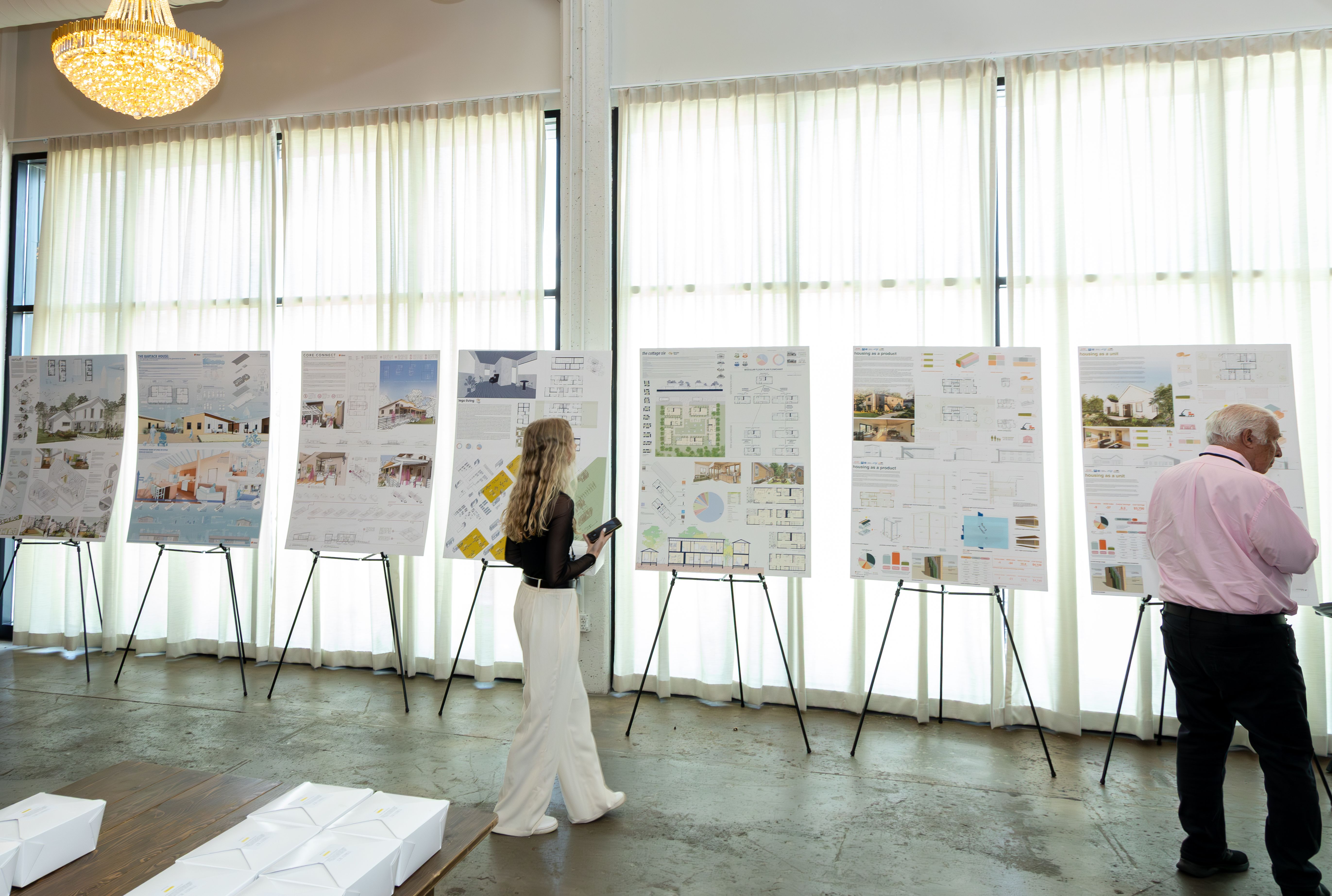 Photograph of multiple detailed architectural display boards set up in an exhibition hall. Titles like 'PHASE I: EQUITABLE HOUSING SOLUTION' are visible, covered in floor plans, sections, and technical diagrams. Attendees in the background are viewing the work.
