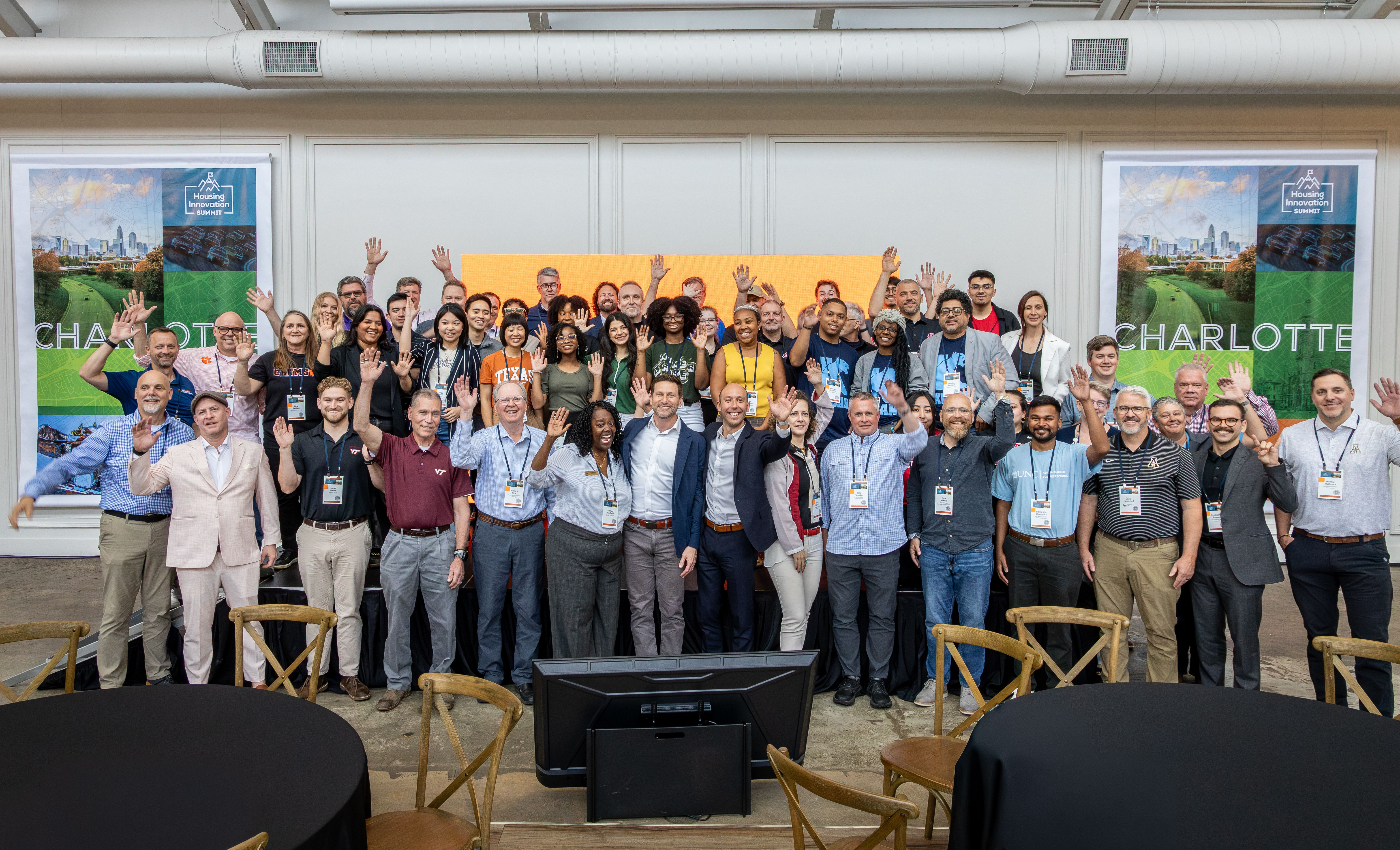 A wide-angle group photograph taken on a stage, featuring the four core team members along with diverse members from other qualifying teams and key leadership figures. Large event banners are overhead in a celebratory atmosphere.